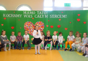 Dziewczynka trzyma w ręku mikrofon, recytuje wiersz, w tle siedzą dzieci w kręgu.